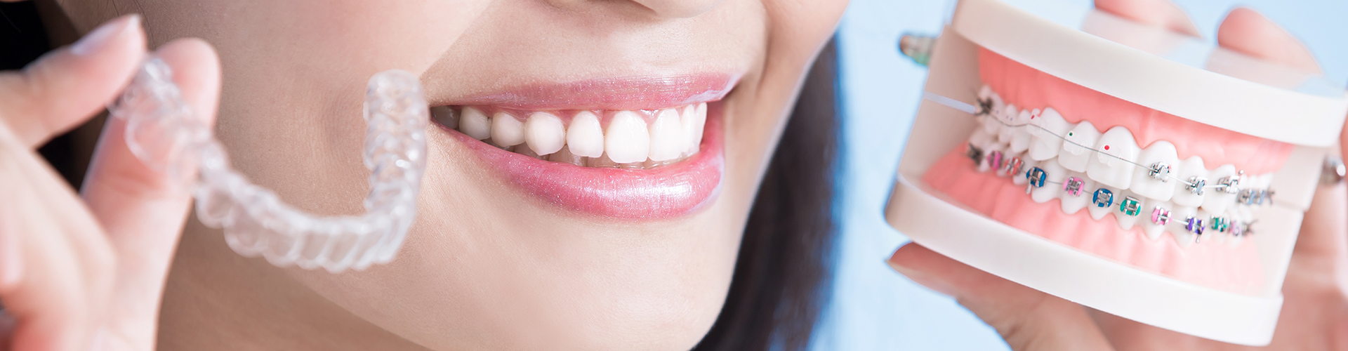 smiling woman holding clear aligner in one hand and braces model in the other